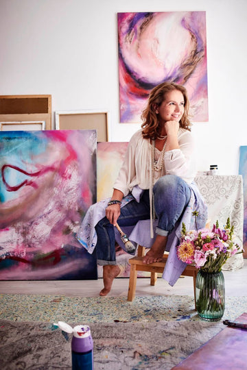A woman artist sitting on a small chair in her studio amongst her colorful pink paintings and a bouquet of lowers in a vase.