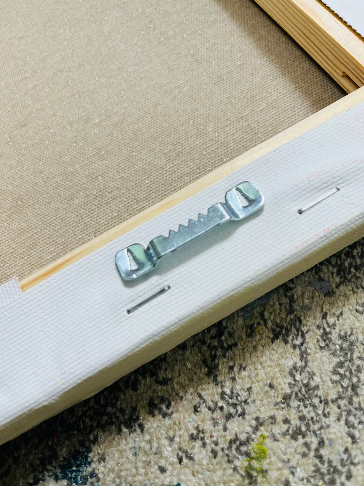 Close-up of the back corner of a canvas print showing the white canvas edge, metal hanging pins, and wooden stretcher bars over a textured carpet.