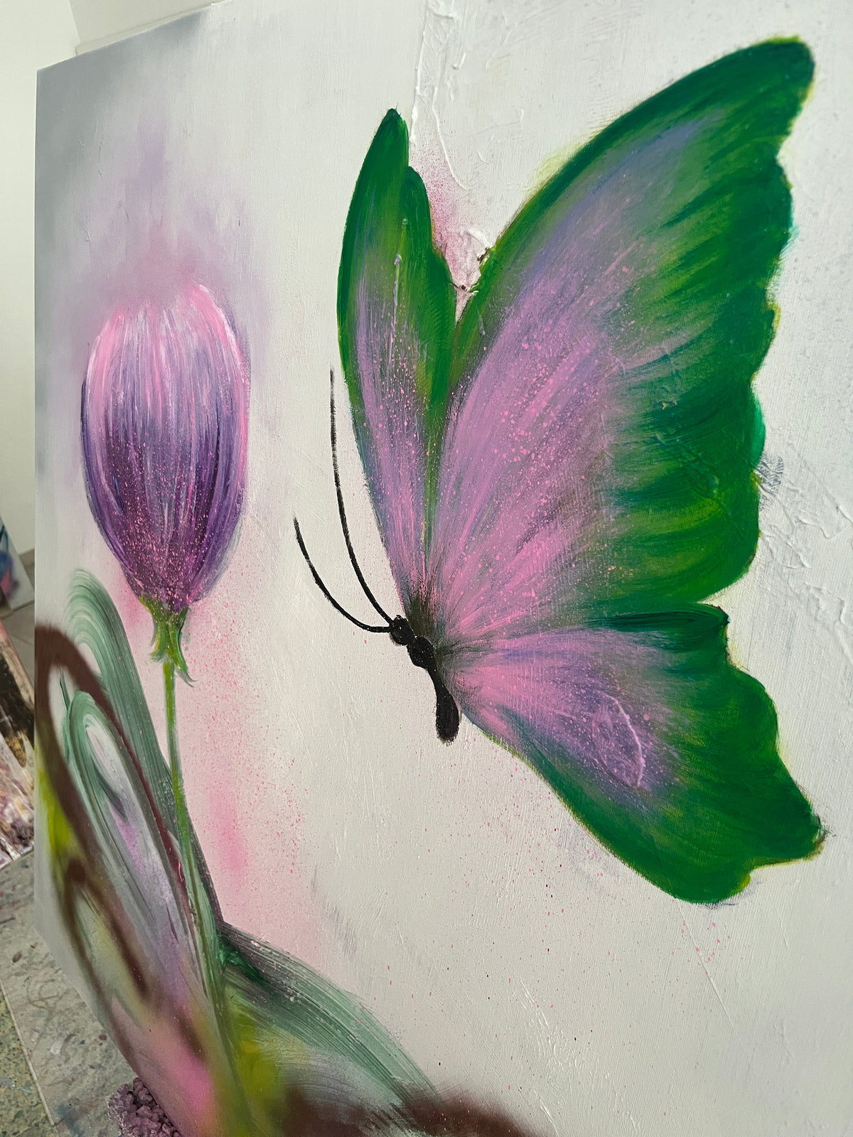 A close-up of a painted artwork featuring a butterfly with green and pink wings next to a purple flower bud on a textured off-white background.