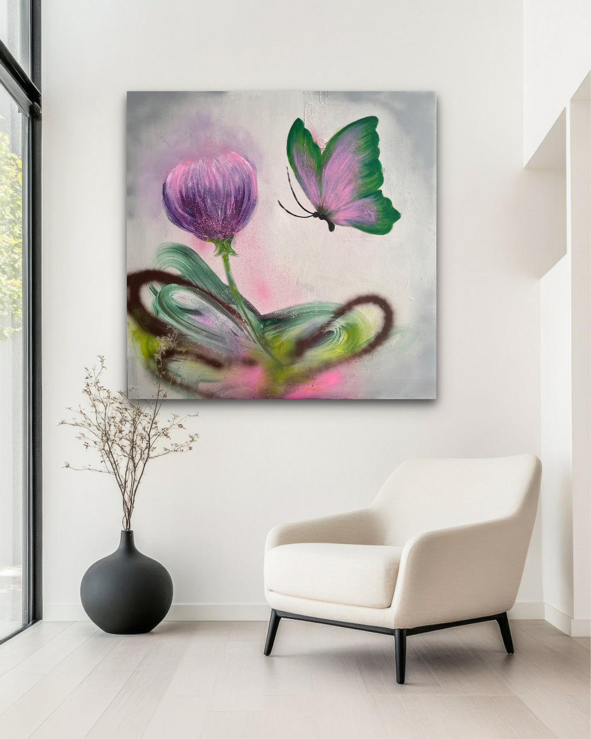 A square wall art print or canvas hanging above a white armchair in a bright interior. The artwork depicts a purple flower, a green-and-pink butterfly, and abstract swirling brushstrokes in pink, green, gray, and brown.