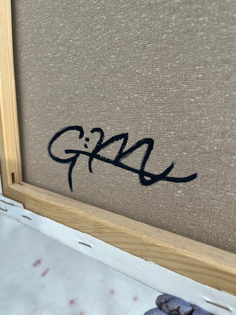 Close-up of a textured beige canvas or fabric panel with a black stylized signature or lettering across the center, bordered by a light wood frame and white edge material.